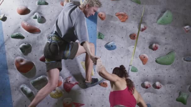 Deportiva y atlético escalar la pared de escalada en el gimnasio. El hombre da una mano amiga, ayuda a levantarse. Utiliza una cuerda de seguridad en la pared de cantos rodados — Vídeos de Stock