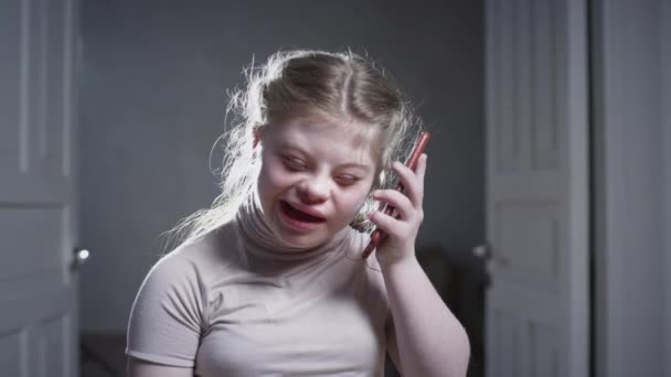 Дівчина з синдромом Дауна використовує смартфон вдома. Дзвонить, розмовляє по телефону, посміхається. Людина з обмеженими можливостями вдома. Життя з інвалідністю — стокове відео