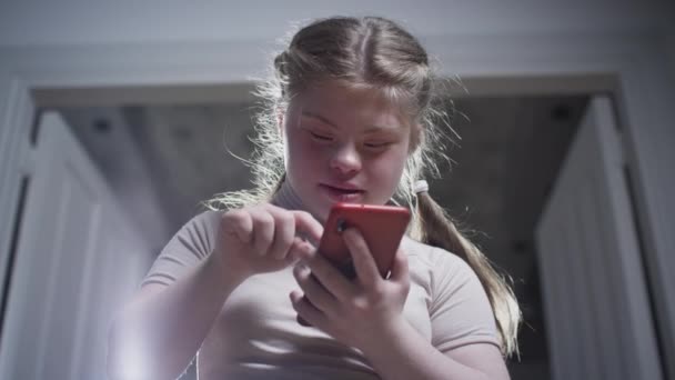 Egy Down-kóros lánynak okostelefonja van. Üzenetet ír, képeket néz, közösségi média alkalmazást használ. Fogyatékkal élő személy otthon — Stock videók
