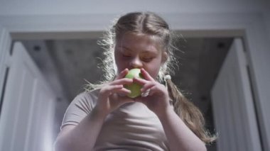 A girl with down syndrome is biting a green apple. Healthy food. Disabled person at home. Life with a disability