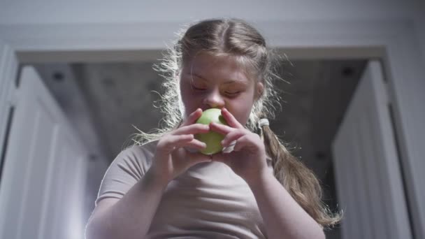 Ένα κορίτσι με σύνδρομο Down δαγκώνει ένα πράσινο μήλο. Υγιεινή τροφή. Αναπηρία στο σπίτι. Ζωή με αναπηρία — Αρχείο Βίντεο
