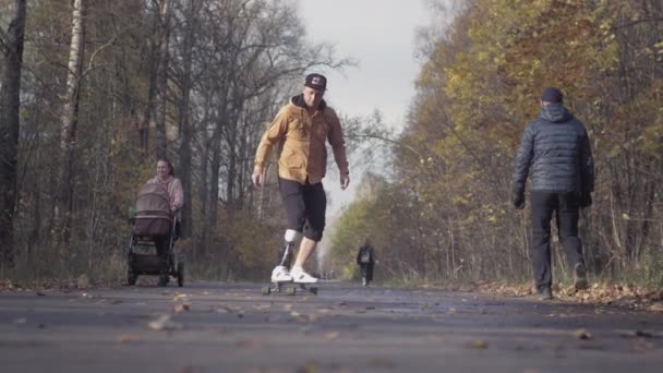 Ein junger Mann mit einer bionischen Beinprothese aus Metall fährt im herbstlichen Wald auf einem Skateboard. Ein künstliches Bein schiebt sich auf einem Skateboard vom Asphalt — Stockvideo