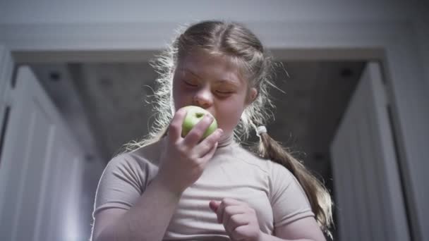 A girl with down syndrome is biting a green apple. Healthy food. Disabled person at home. Life with a disability — Stockvideo