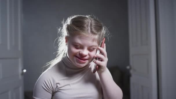 A girl with down syndrome uses a smartphone at home. Makes a call, talks on the phone, smiles. Disabled person at home. Life with a disability — Stock Video