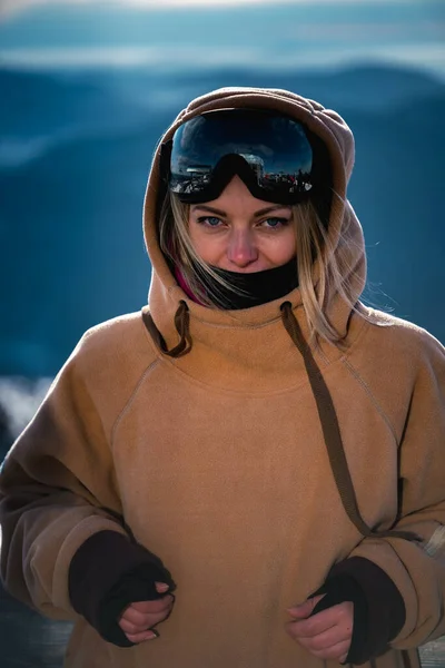 Portret Piękna dziewczyna w ośrodku narciarskim na tle śnieżnych gór. Jest ubrana w zimowe ubrania do snowboardu i gogli narciarskich — Zdjęcie stockowe