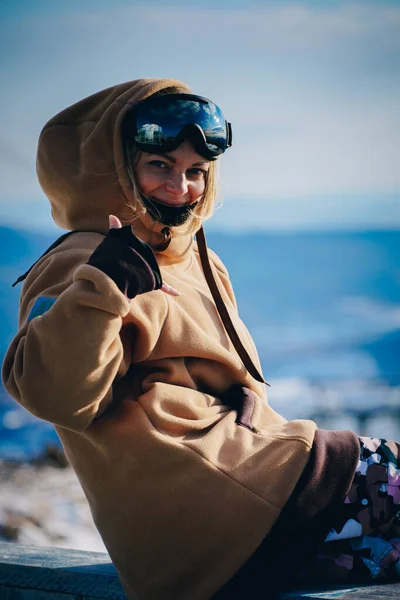 Portret Piękna młoda dziewczyna w ośrodku narciarskim na tle śnieżnych gór. Jest ubrana w zimowe ubrania do snowboardu i gogli — Zdjęcie stockowe