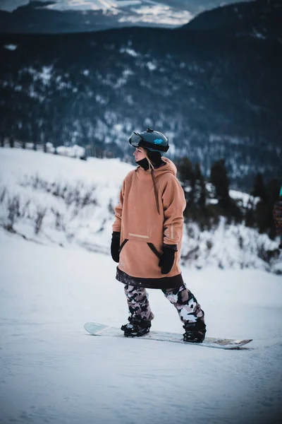 Retrato Menina bonita em uma estância de esqui contra o pano de fundo de montanhas nevadas. Ela está vestida com roupas de inverno para snowboard e óculos — Fotografia de Stock