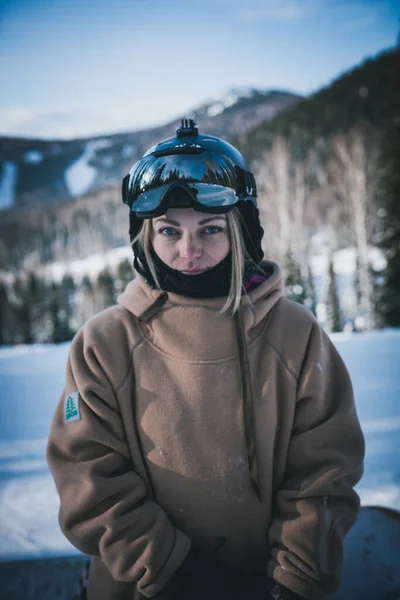 Retrato Menina bonita em uma estância de esqui contra o pano de fundo de montanhas nevadas. Ela está vestida com roupas de inverno para snowboard e óculos — Fotografia de Stock