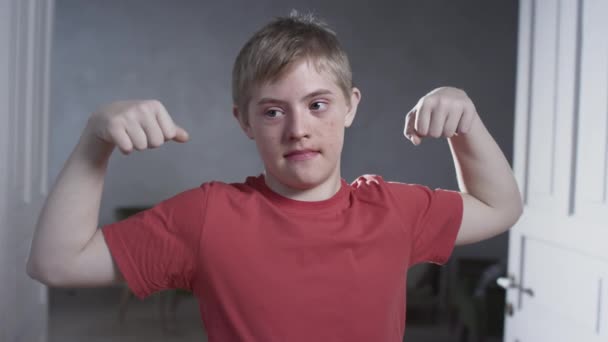 Retrato de un tipo con síndrome de Down. El niño demuestra músculos y bíceps en casa. Entrenamientos en casa y deportes. Una persona con necesidades especiales — Vídeo de stock