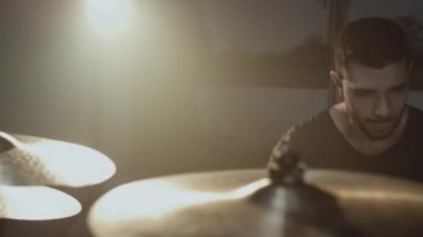 Genç ve çekici bir erkek davulcu bateri setinde bir sopayla çalıyor. Metal zillere vuruyor. Ses ve şarkılar için ev yapımı prova stüdyosu.