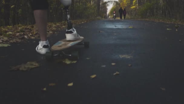 Een jongeman met een metalen kunstbeen rijdt op een skateboard in een herfstpark. Ga sporten met een kunstbeen — Stockvideo