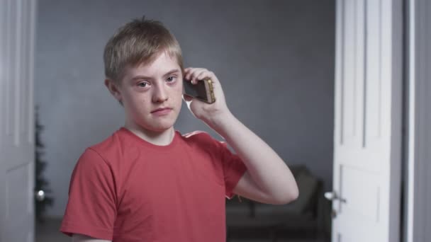 Ritratto di un ragazzo con sindrome di Down. Il bambino tiene uno smartphone con la mano, sta parlando al telefono. Disturbo genetico cromosomico in un bambino. — Video Stock
