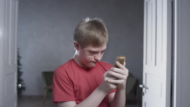 Portrait of a boy with Down Syndrome. The kid holds a smartphone with his hands, plays mobile games in the application. A person with special needs — Stock Video
