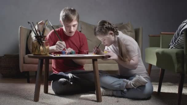 Två barn med Downs syndrom. En pojke och en flicka ritar med färger och en pensel. Kommunikation för barn med funktionshinder. En person med särskilda behov — Stockvideo