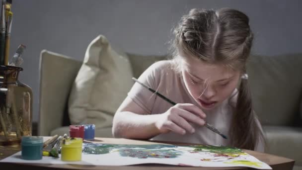 Ritratto di una ragazza con sindrome di Down. La bambina disegna con vernici e un pennello a casa. Crea arte e creatività. Una persona con bisogni speciali — Video Stock