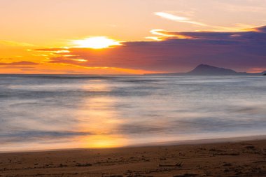 Gün doğumu veya gün batımı sahilde, gökyüzünde bulutlar ve ufukta dağlarla.