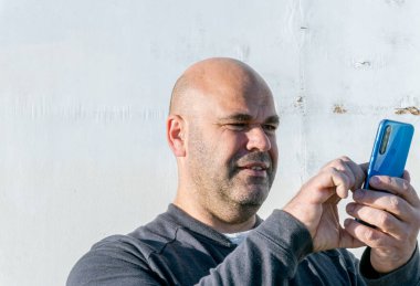 Orta yaşlı beyaz adam portresi, biraz sakallı, tıraş olmuş saçı, akıllı telefon ekranı, beyaz ahşap arka planda..