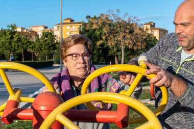 Erkek spor koçu, yaşlı bir kadının gün batımında biyo-sağlıklı bir parkta tekerlekleri hareket ettirmesine yardım ediyor. Biyo-sağlıklı park, gün batımında.