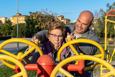 Erkek spor koçu, yaşlı bir kadının gün batımında biyo-sağlıklı bir parkta tekerlekleri hareket ettirmesine yardım ediyor. Biyo-sağlıklı park, gün batımında.