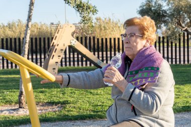 Yaşlı kadın sporcu, güneş batarken, spor bisikletinin üstünde biyo-sağlıklı bir parkta şişeden su içiyor.