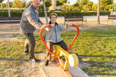 Erkek antrenör, yaşlı kadının gün batımında biyo-sağlıklı bir parkta makinenin üzerinde egzersiz yapmasına yardım eder.