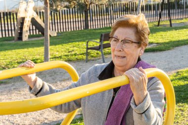 Gün batımında parkta biyo-sağlıklı makinelerle egzersiz yapan yaşlı bir kadın..