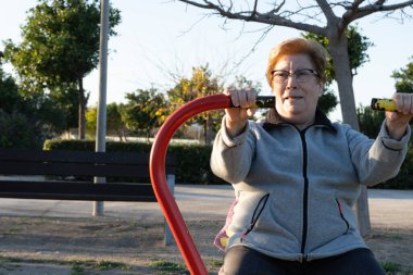 Gün batımında parkta biyo-sağlıklı makinelerle egzersiz yapan yaşlı bir kadın..