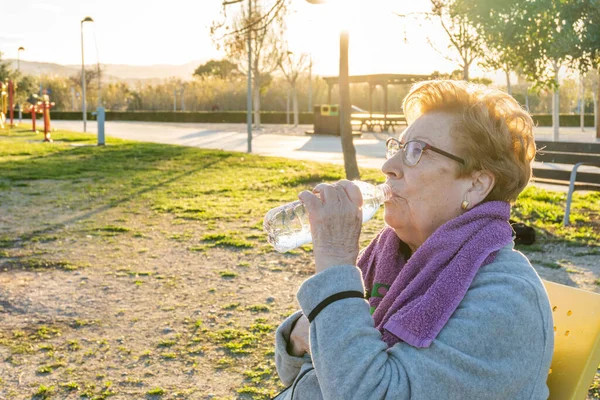 Yaşlı kadın gün batımında parkta egzersiz yaptıktan sonra şişeden su içiyor.