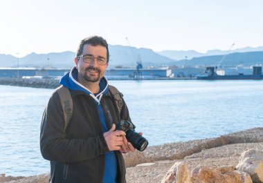 Sırıtkan orta yaşlı profesyonel bir fotoğrafçının portresi, elinde profesyonel bir refleks kamerayla, arka planda bir porto ile.