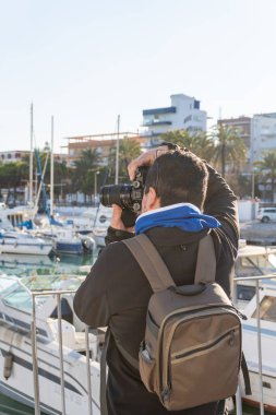 Orta yaşlı bir adamın günbatımında bir limanda profesyonel bir refleks kamerayla fotoğraf çekerken görüntüsü.