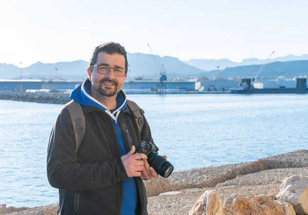 Sırıtkan orta yaşlı profesyonel bir fotoğrafçının portresi, elinde profesyonel bir refleks kamerayla, arka planda bir porto ile.