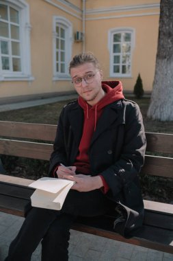 Çocuk parkta bankta oturuyor ve deftere bir şeyler yazıyor.