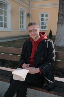 Çocuk parkta bankta oturuyor ve deftere bir şeyler yazıyor.