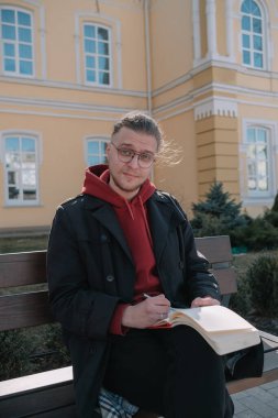 Çocuk parkta bankta oturuyor ve deftere bir şeyler yazıyor.