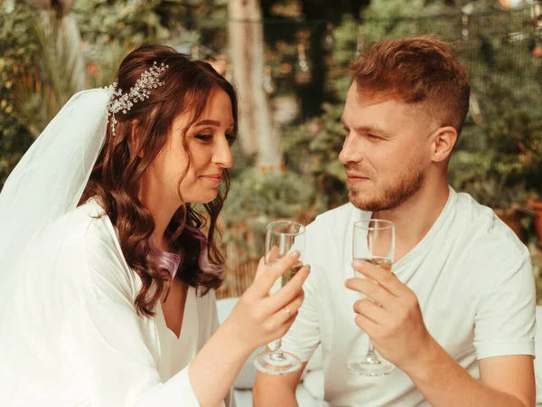 Şık, güler yüzlü damat ve beyaz elbiseli, kıvırcık saçlı tatlı sarışın gelin halının üzerinde şampanya kadehleriyle oturuyorlar. tahıl etkisi — Stok fotoğraf