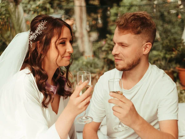 Şık, güler yüzlü damat ve beyaz elbiseli, kıvırcık saçlı tatlı sarışın gelin halının üzerinde şampanya kadehleriyle oturuyorlar. tahıl etkisi — Stok fotoğraf