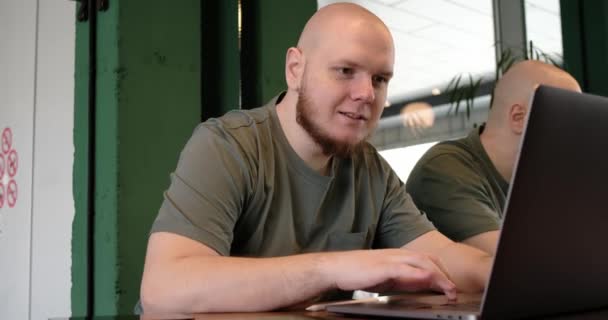 Hombre feliz sin afeitar usando el ordenador portátil mientras está sentado en la cafetería — Vídeos de Stock