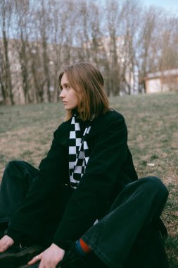 Siyah ceketli kadın parkta yerde oturuyor.