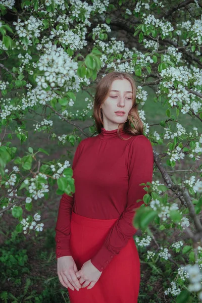 Kırmızılı sarışın bir kız çiçek açan ağaçların bahçesinde doğayla birleşiyor.