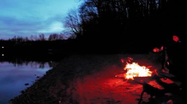 Yalnız bir adam gölün kıyısındaki orman ateşinin yanında oturuyor. 