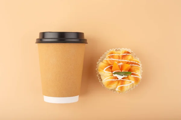 Top view of coffee or tea in brown cardboard cup with pastry against beige background. Concept of hot drinks, take away food, small breaks or cheat meal