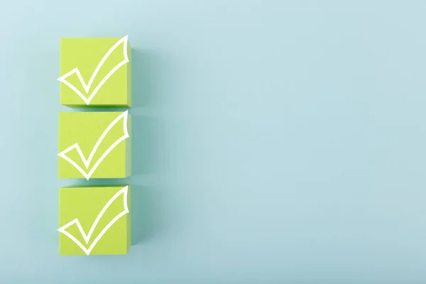 Three white checkmarks on green cubes against bright pastel blue background with copy space — Foto Stock