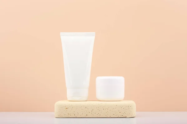 White cosmetic tubes with scrub and mask for face or hand skin on gypsum podium on white table against beige background. Imagem De Stock