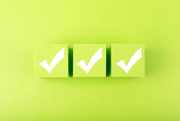 Three white checkmarks on green toy cubes in a row against bright green background. Checklist concept Imagem De Stock