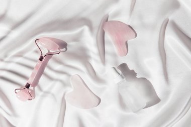 Evde cilt bakımı ve spa için makyaj seti. Yüz silindiri ve gua sha masaj aletleri, beyaz ipek arka planda bir şişe gerekli yağ ya da serum. Doğal tedavi konsepti. Üst Manzara. Düz yatıyordu