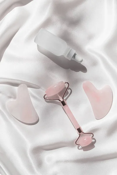 Evde cilt bakımı ve spa için makyaj seti. Yüz silindiri ve gua sha masaj aletleri, beyaz ipekten arka planda bir şişe gerekli yağ ya da serum. Doğal tedavi konsepti. Üst Manzara. Düz yatıyordu