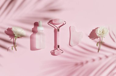 Yüz silindirinden, gua sha masajcıdan, pembe pastel renkli, beyaz gül tomurcukları ve gölgelerle aydınlatılmış bir şişe kozmetik serumdan oluşan yaratıcı bir kompozisyon. Doğal tedavi konsepti. Üst Manzara. Düz yatıyordu