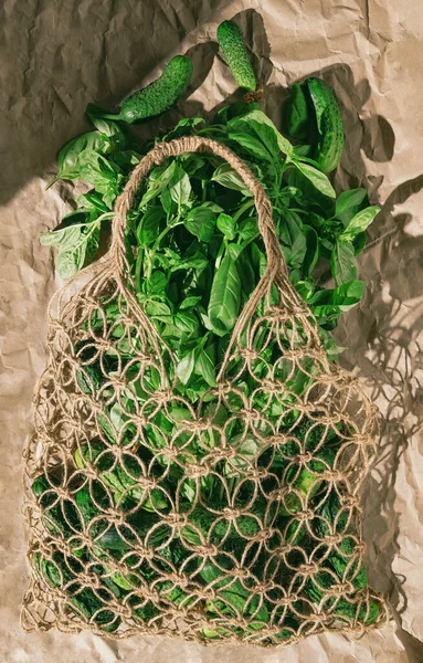 Creative layout made of fresh basil and cucumbers in mesh bag on sunlit craft paper background with shadow. Healthy food ingredient concept. Top view. Flat lay