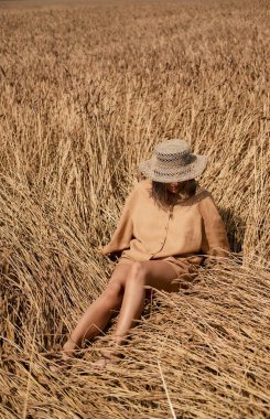 Keten giysiler içinde çıplak ayaklı genç bir kadın ve arka planda kuru otlardan oluşan bir şapka. Şapkalı romantik kız çayırda oturuyor, doğada tek başına..