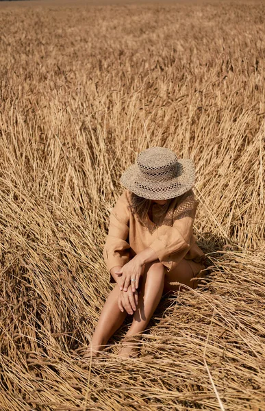 Young woman barefoot in linen clothes and a hat on a background of dry grass. Romantic girl in a hat sits on a meadow, relaxed alone in nature. — Stock Photo, Image